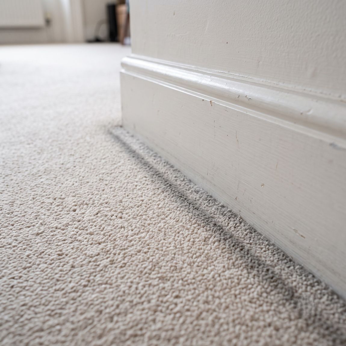 A subtle but clearly visible greyish filtration line runs continuously along the very edge of a light beige, off-white or pale cream fitted carpet in a standard, lived-in Central London apartment